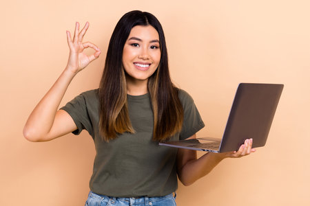 Photo of attractive malaysian girl toothy smile demonstrate okey symbol hold wireless netbook isolated on beige color backgroundの写真素材