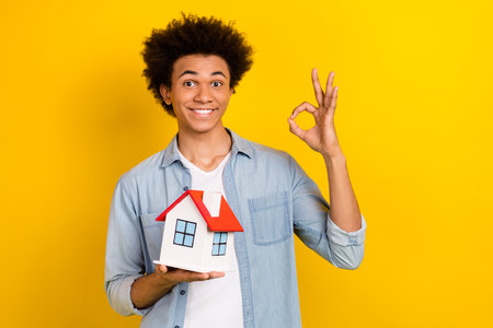 Photo of funky attractive man wear jeans shirt showing okey sign holding little house isolated yellow color backgroundの写真素材