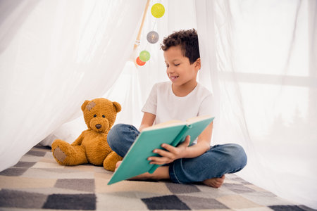 Photo of smart schoolkid reading book telling story teddy bear in comfort homemade tent indoors in preschool playroomの写真素材