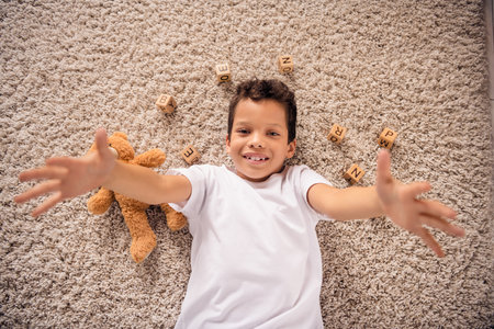 Portrait of nice cheerful cute little boy lying floor soft carpet stretch hands wear white shirt light modern room design apartmentの写真素材