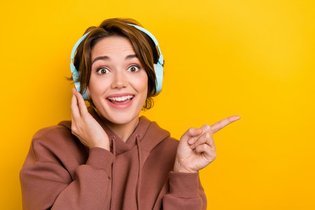 Portrait of charming young girl wear brown pullover pointing finger empty space new apple earphones isolated on yellow color backgroundの写真素材