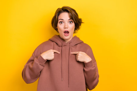 Photo of confused woman with short hairdo wear brown hoodie directing at herself staring open mouth isolated on yellow color backgroundの写真素材