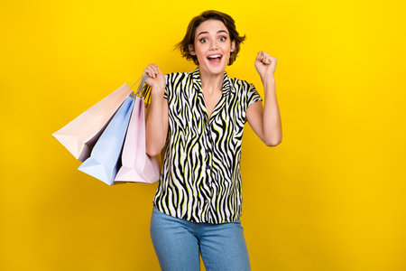 Portrait of impressed woman with short hairstyle wear striped blouse raising fist up hold shopping bag isolated on yellow color backgroundの写真素材