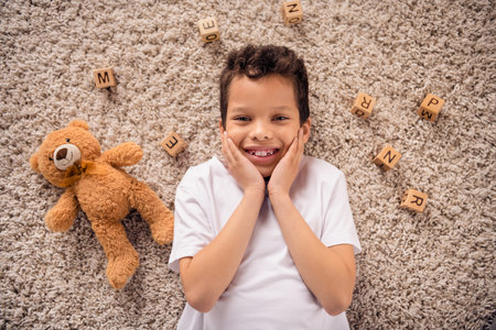 Portrait of charming happy small boy touch cheeks lying soft floor wear white shirt stylish bright child room interior carpet decorationの写真素材