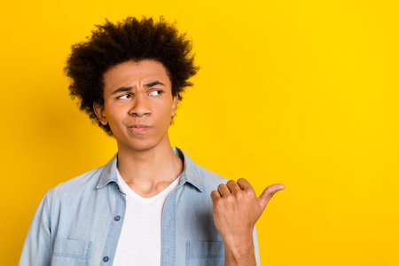 Photo of unsure uncertain man wear jeans shirt pointing thumb looking empty space isolated yellow color backgroundの写真素材