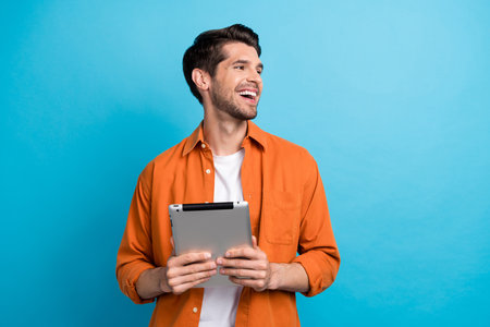 Photo of positive excited guy wear orange shirt texting modern device looking empty space isolated blue color backgroundの写真素材