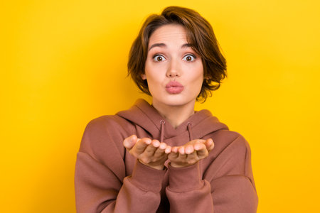 Portrait of young flirting woman wear brown sweatshirt send you hands air kiss girlfriend love story isolated on yellow color backgroundの写真素材