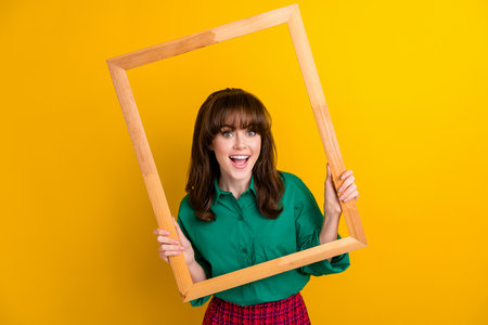 Photo of excited funky woman dressed green shirt tacking photo wooden frame isolated yellow color...