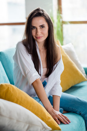 Vertical photo of pretty positive young lady sitting comfy couch wear shirt bare shoulder bright apartment indoorsの写真素材