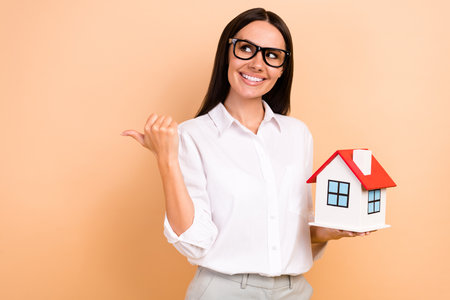 Portrait of beautiful charming girl arm hold little house look direct finger empty space isolated on beige color backgroundの写真素材