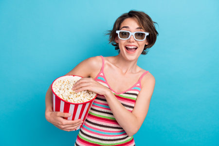 Photo of overjoyed cheerful girl hold big popcorn box enjoy watching new movie 3d glasses isolated on blue color backgroundの写真素材