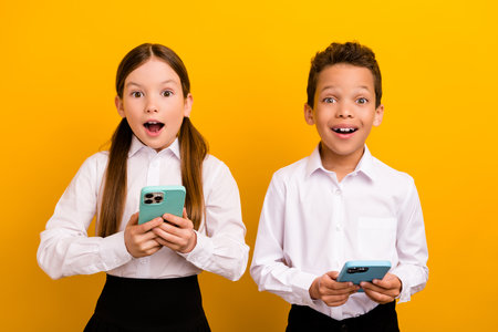 Photo of sweet impressed preteen schoolchildren wear white shirts communicating modern gadgets isolated yellow color backgroundの写真素材