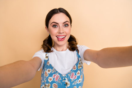 Photo of flirty cute girl dressed flower print overall recording video vlog showing tongue isolated beige color backgroundの写真素材