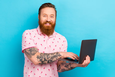 Photo of positive cheerful intelligent man with redhair beard wear pink t-shirt typing email on laptop isolated on blue color backgroundの写真素材
