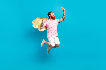 Full size portrait of excited handsome guy jump hold boutique bags smart phone make selfie portrait isolated on blue color backgroundの写真素材