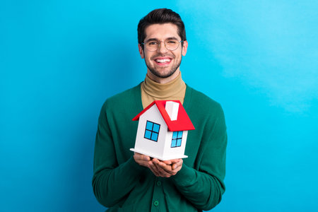 Portrait of successful positive man real estate sales manager toothy smile hands hold small house isolated on blue color backgroundの写真素材