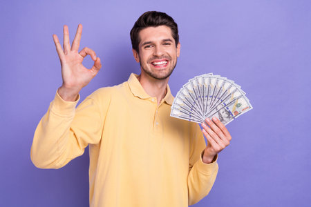 Photo of attractive positive guy toothy smile arm hold dollar banknotes bills demonstrate okey symbol isolated on violet color backgroundの写真素材