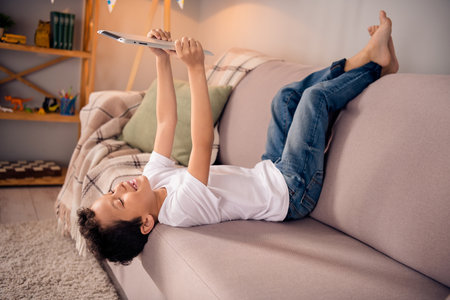Portrait of adorable small schoolboy lying upside down sofa tablet wear white shirt modern interior living room playroom daycare indoorsの写真素材