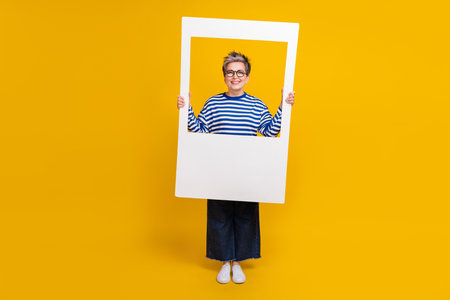 Full length photo of good mood senior lady wear stylish clothes hold cadre picture photography isolated on yellow color backgroundの写真素材