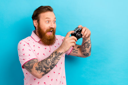 Portrait of impressed astonished man with cool tattoo wear flamingo t-shirt hold camera take photo isolated on blue color backgroundの写真素材