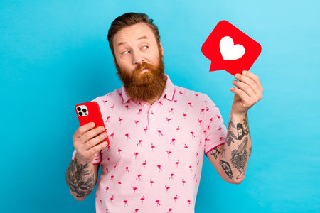 Portrait of interested man with cool tattoo wear flamingo t-shirt hold smartphone look at like in hand isolated on blue color backgroundの写真素材