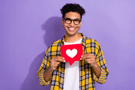 Photo of pretty cheerful guy dressed checkered yellow shirt eyewear holding heart like card isolated...