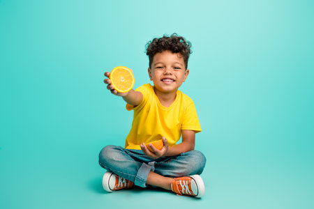 Full size photo of adorable small boy sit floor hold orange slice wear trendy yellow outfit isolated on aquamarine color backgroundの写真素材