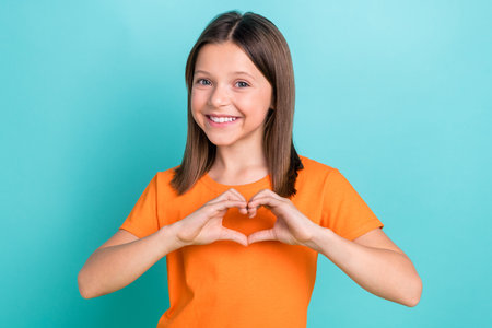 Portrait of toothy beaming schoolgirl with straight hair dressed orange clothes show heart on chest isolated on turquoise color backgroundの写真素材