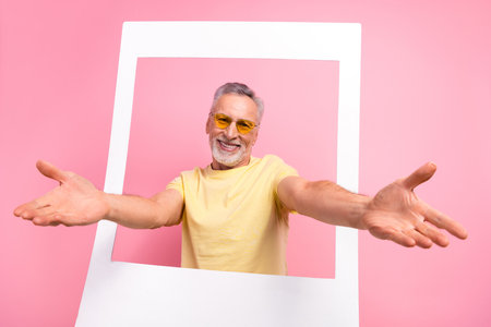 Photo of friendly cheerful man with beard dressed yellow t-shirt in sunglass stretching arms from frame isolated on pink color backgroundの写真素材