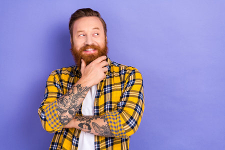 Portrait of minded creative cheerful man hand touch beard chin look interested empty space isolated on purple color backgroundの写真素材