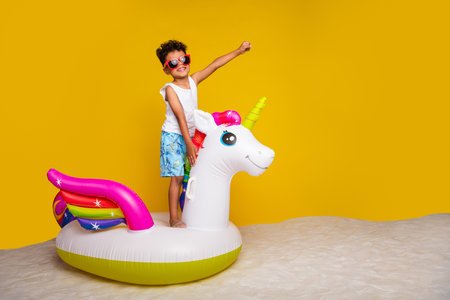 Full length photo of cheerful cool small boy wear white shirt riding floating unicorn empty space isolated yellow color backgroundの写真素材