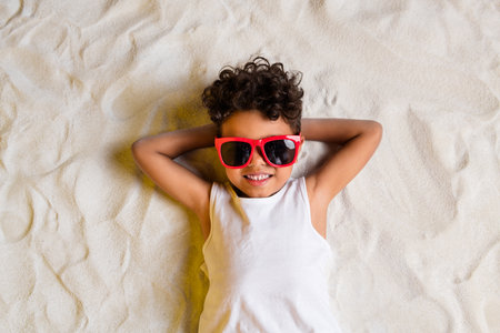 High angle view photo of charming cheerful little boy wear white shirt dark spectacles lying arms behind head outside sea beachの写真素材