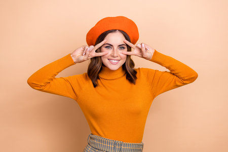 Portrait of cheerful gorgeous lady toothy smile arms fingers show v-sign near eyes isolated on beige color backgroundの写真素材