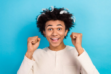 Photo portrait of cute young guy raise fists feathers afro hair dressed white pajama outfit isolated...