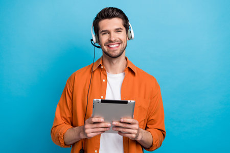 Portrait of handsome positive guy toothy smile hold use tablet wired headset communicate clients isolated on blue color backgroundの写真素材