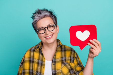 Photo portrait of charming grandmother hold heart icon influencer wear trendy plaid yellow outfit isolated on cyan color backgroundの写真素材