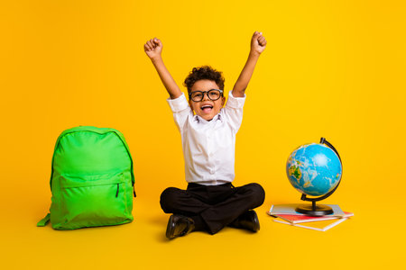 Full body photo of little overjoyed schoolchild sit floor raise fists shout yes world globe rucksack book isolated on yellow color backgroundの写真素材