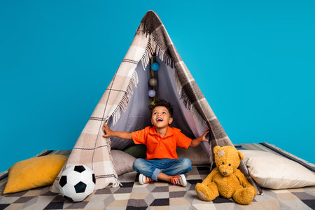 Photo of amazed surprised adorable boy playing with handmade tent isolated on blue color background