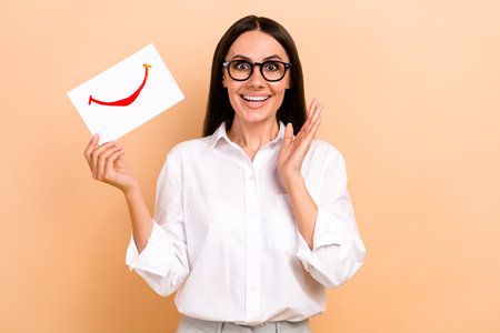 Photo of excited amazed woman dressed white shirt eyewear folding smiling mouth card isolated beige color backgroundの写真素材