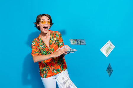 Portrait of astonished wealthy girl arms throw flying dollar banknotes bills isolated on blue color...