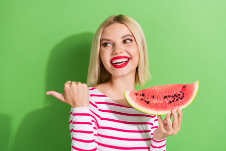 Portrait of funny positive girl dressed striped clothes hold watermelon look directing empty space isolated on green color backgroundの写真素材