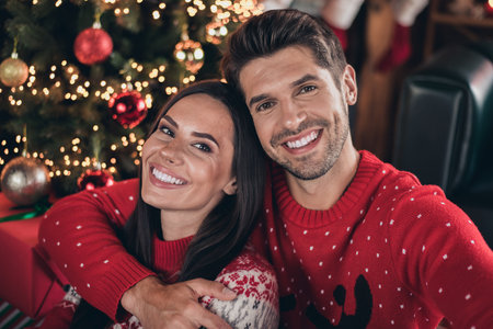 Close up photo of cute pretty smile couple wearing red warm sweaters winter holiday ornament hugs isolated on evergreen tree backgroundの写真素材