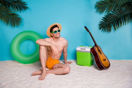 Full length photo of charming dreamy shirtless man straw hat sitting sand looking empty space isolated blue color backgroundの写真素材