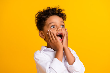 Photo of astonished boy with curly hair dressed white shirt palms on cheekbones staring at empty space isolated on yellow color backgroundの写真素材