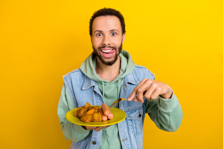 Photo of excited handsome guy tongue lick teeth arm hold fork fried potato sausage plate isolated on yellow color backgroundの写真素材