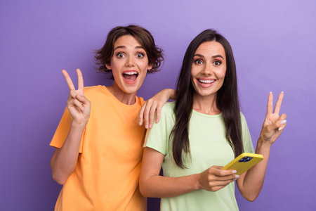 Photo of cute impressed girls dressed t-shirts online communicating modern device showing v-sign...