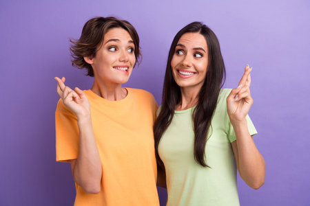 Photo of funky nervous girls dressed t-shirts biting lip fingers crossed isolated purple color backgroundの写真素材