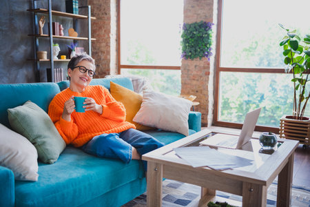 Photo of cheerful positive elderly lady wear orange pullover enjoying coffee watching film modern gadget indoors apartment roomの写真素材