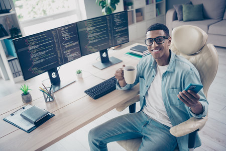 Photo of positive young coder guy browsing phone holding mug with fresh coffee on online meeting...