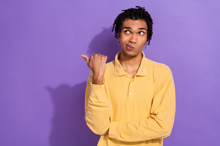Portrait of confused puzzled guy with earrings dressed yellow shirt directing look at empty space isolated on violet color backgroundの写真素材